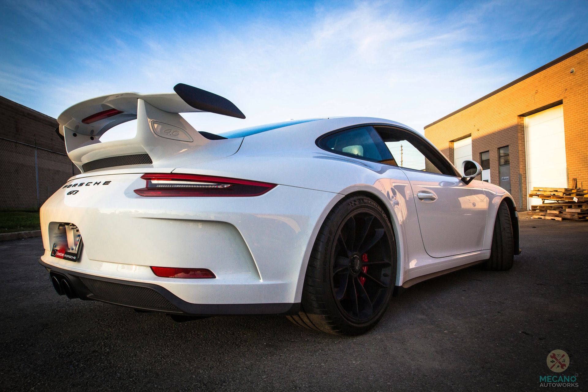 White Porsche 911 GT3 with a large rear wing, black wheels, parked in front of a brick building. | Mecano Autoworks