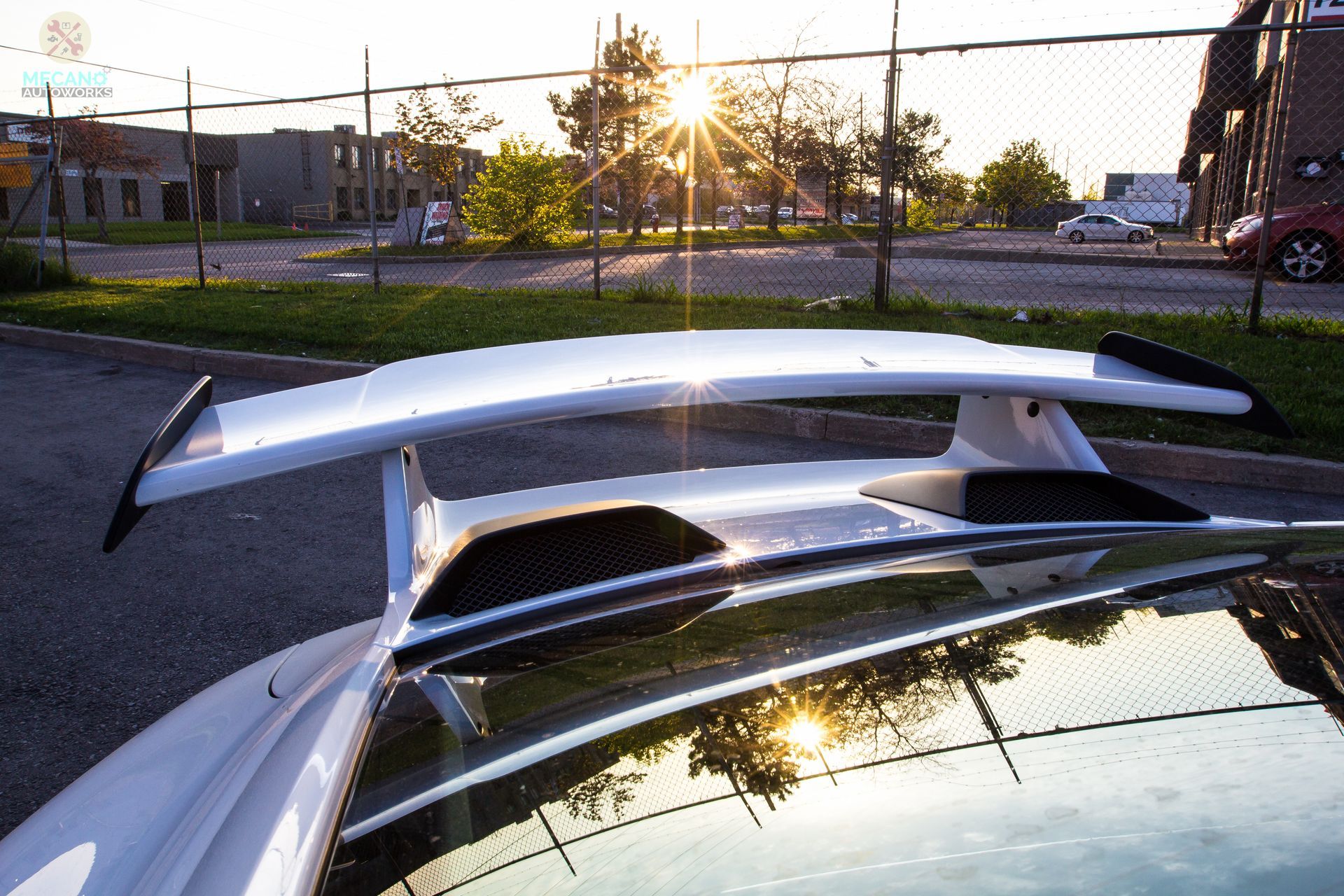 White sports car with large rear wing, sunlight reflecting off the roof, urban setting. | Mecano Autoworks
