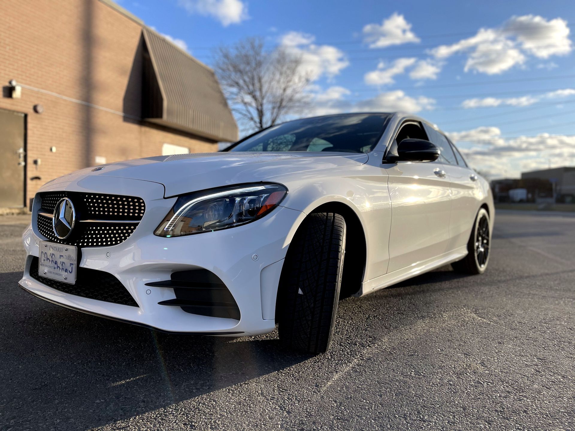 White Mercedes sedan parked on asphalt in front of a brick building under a partly cloudy sky. | Mecano Autoworks