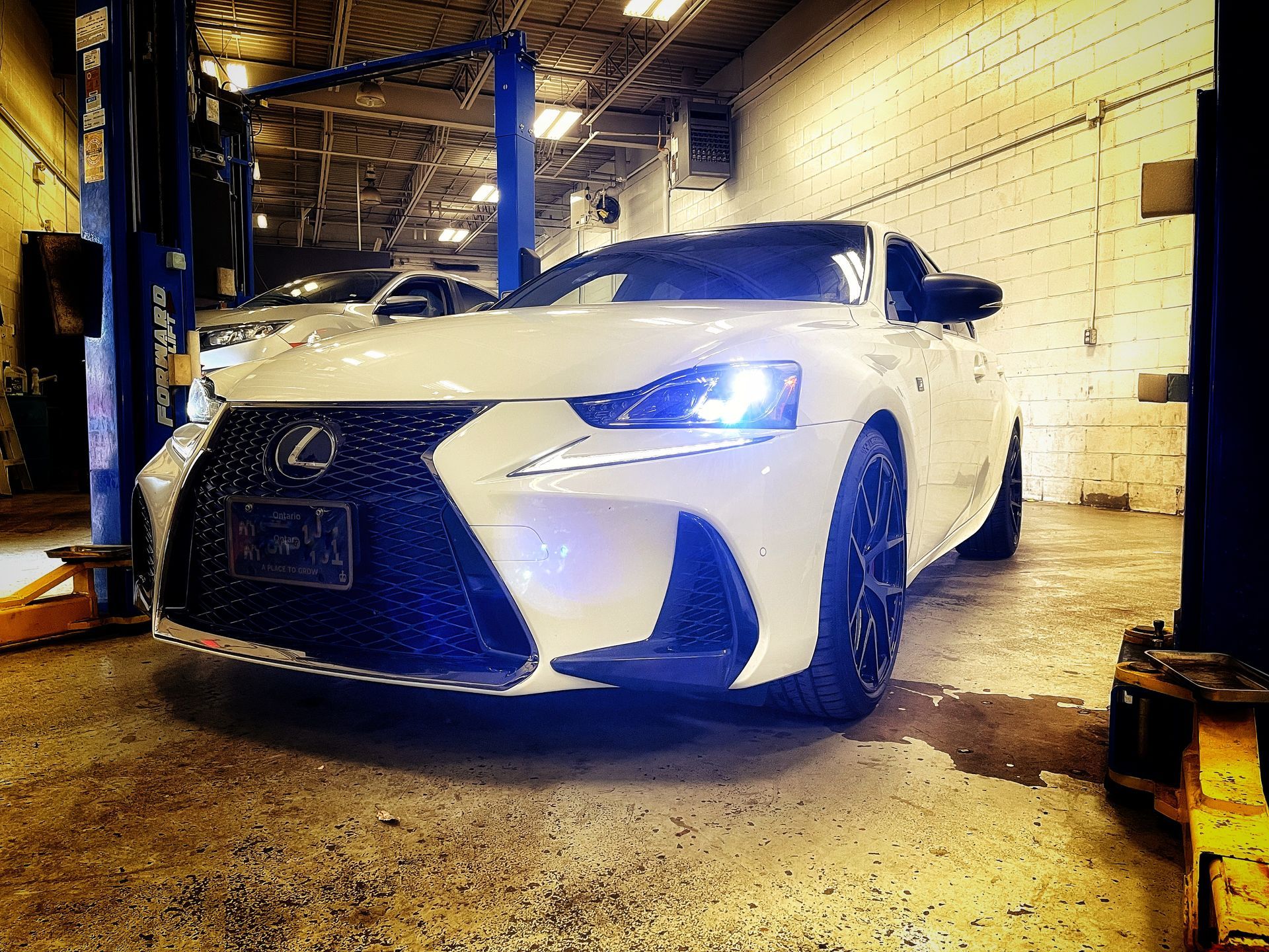 White Lexus sports car in a dimly lit garage with blue headlights, a black grill, and black wheels. | Mecano Autoworks