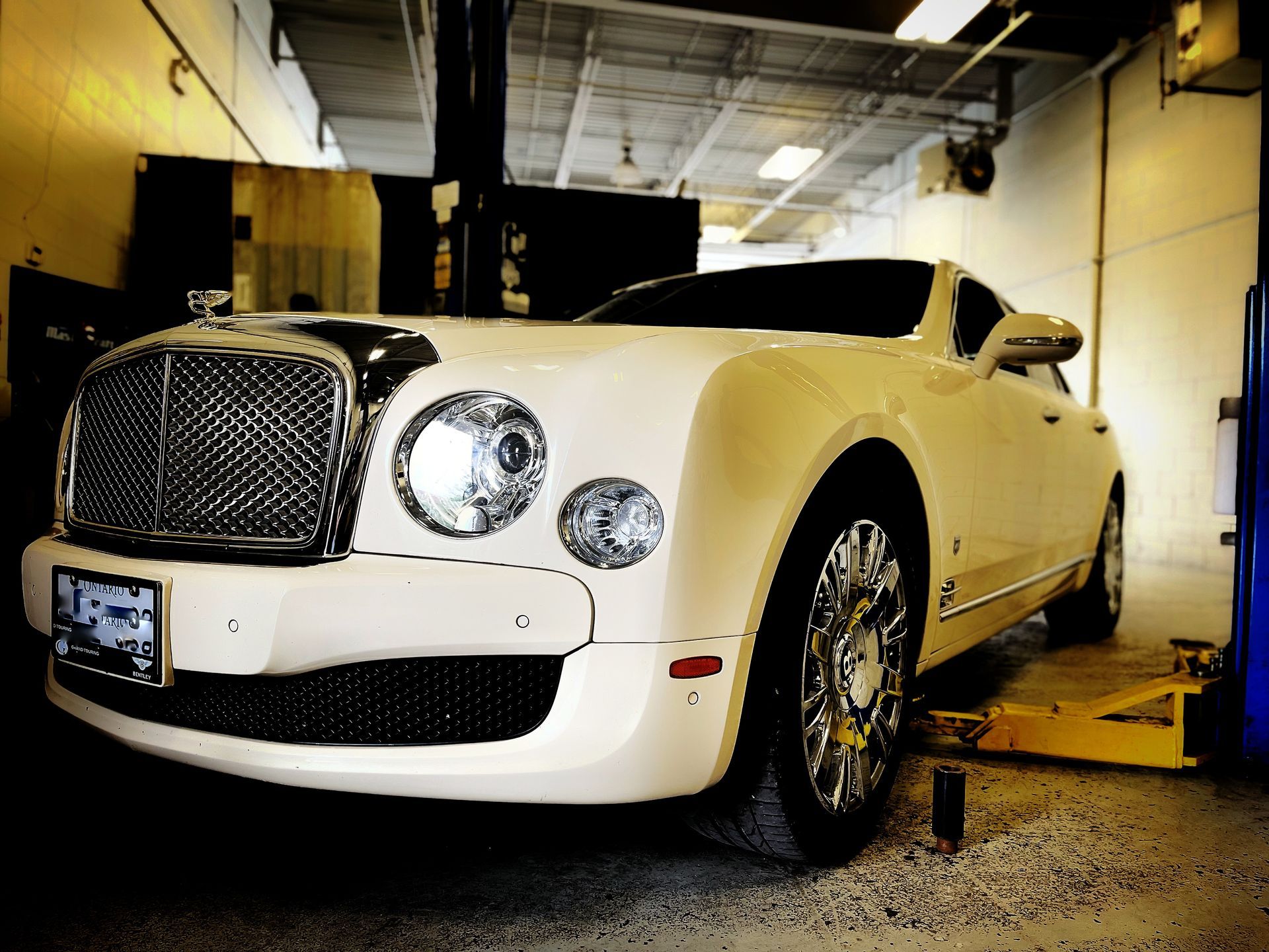 Cream-colored Bentley in a repair shop with its front wheels on a lift, chrome grill, and bright headlights. | Mecano Autoworks