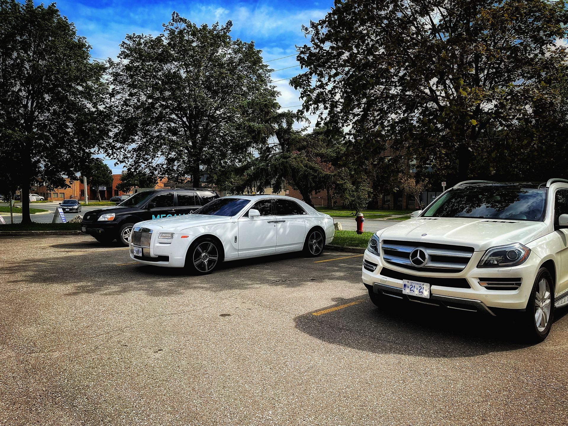 Cars parked in a lot, including a white Rolls Royce and a white Mercedes SUV under trees on a sunny day. | Mecano Autoworks
