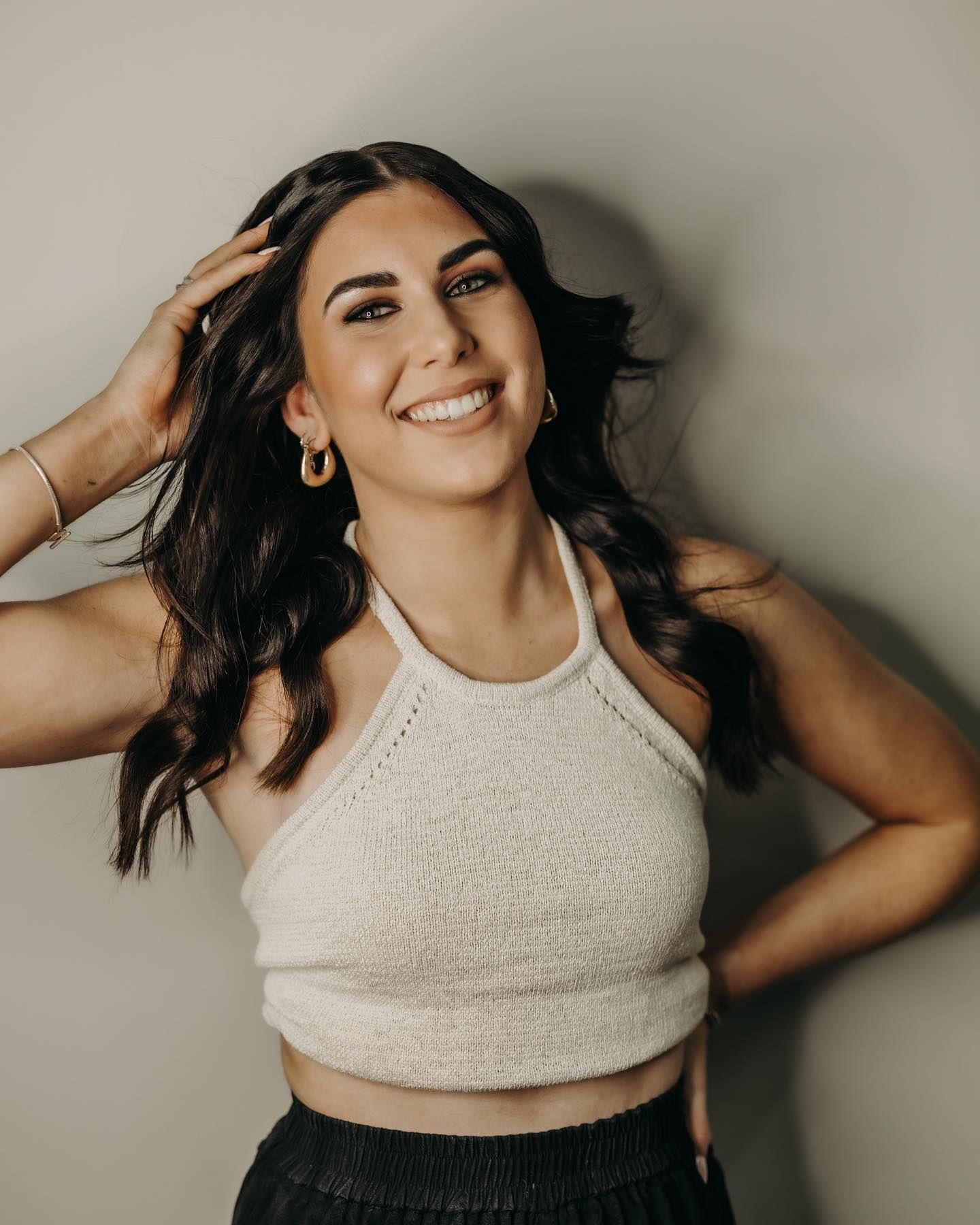 Woman Smiles, Hand in Hair, Wearing White Halter Top, Black Pants, Hoop Earrings — EDISON Hair Collective In Mackay, QLD