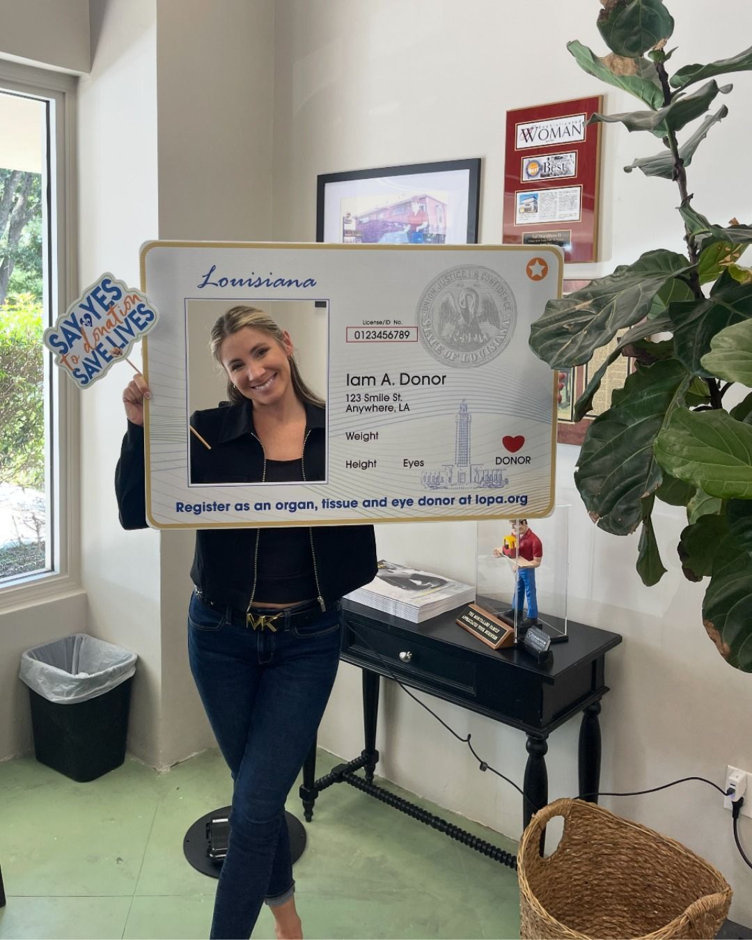 Woman holding a large Louisiana driver's license cutout, smiling, inside a building.