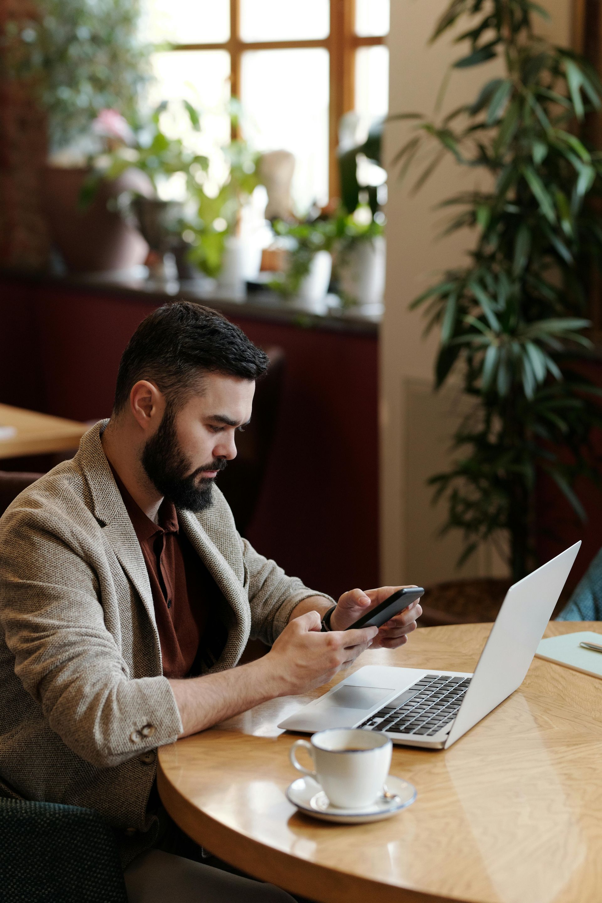 Man using phone and laptop at table, coffee cup, plants and window in background.