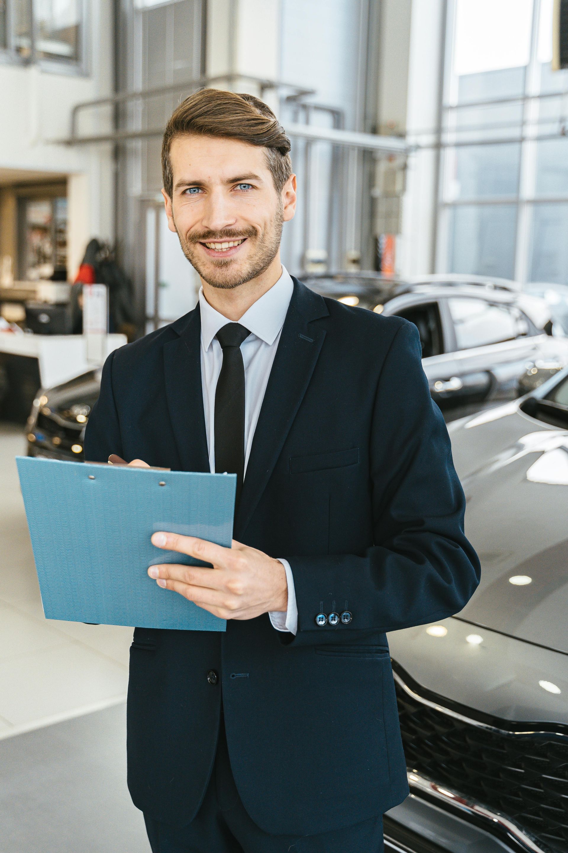 Car salesman holding a clipboard, smiling in a dealership.