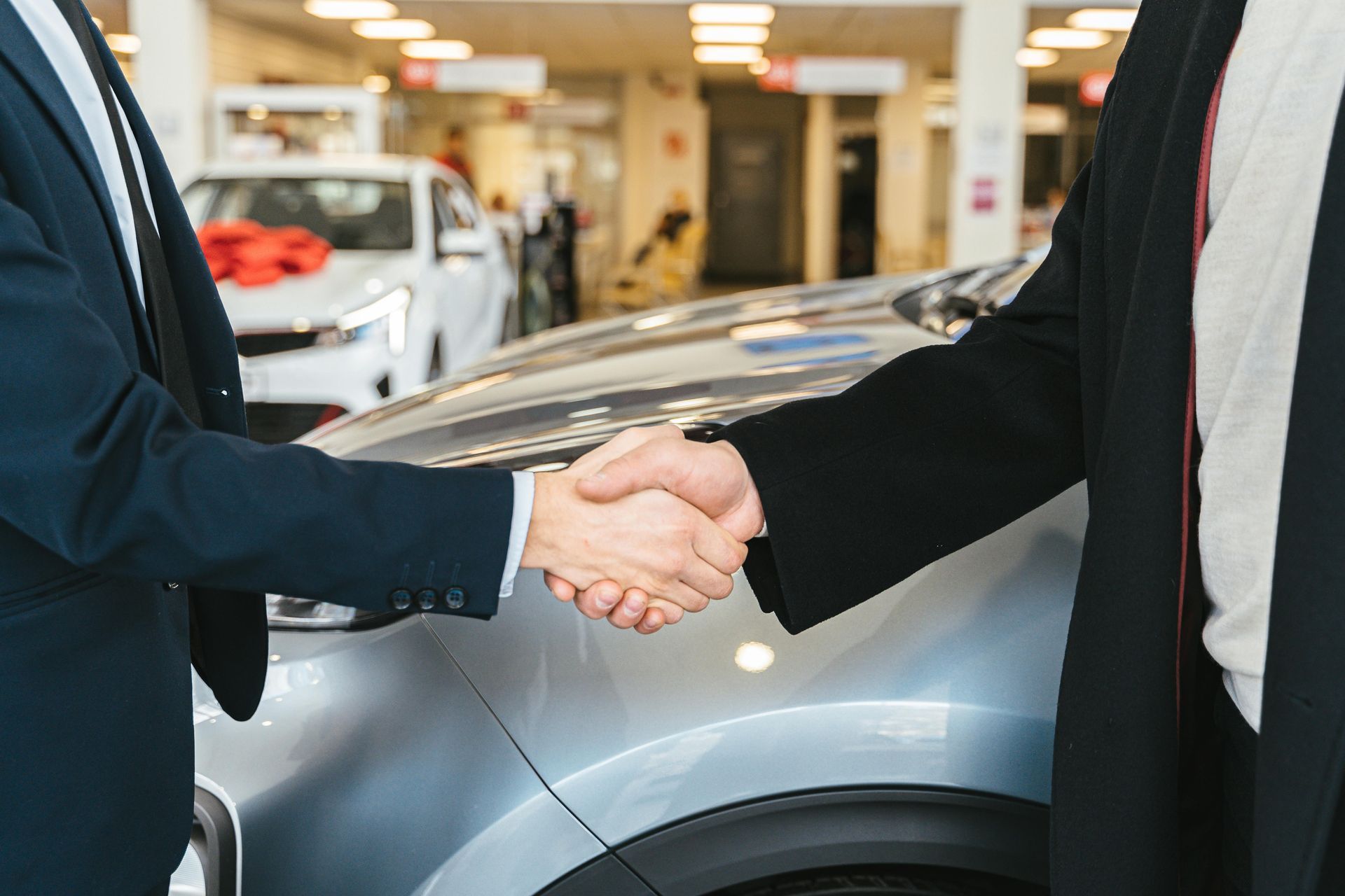 Two men shaking hands in a car dealership, a new car in the background.