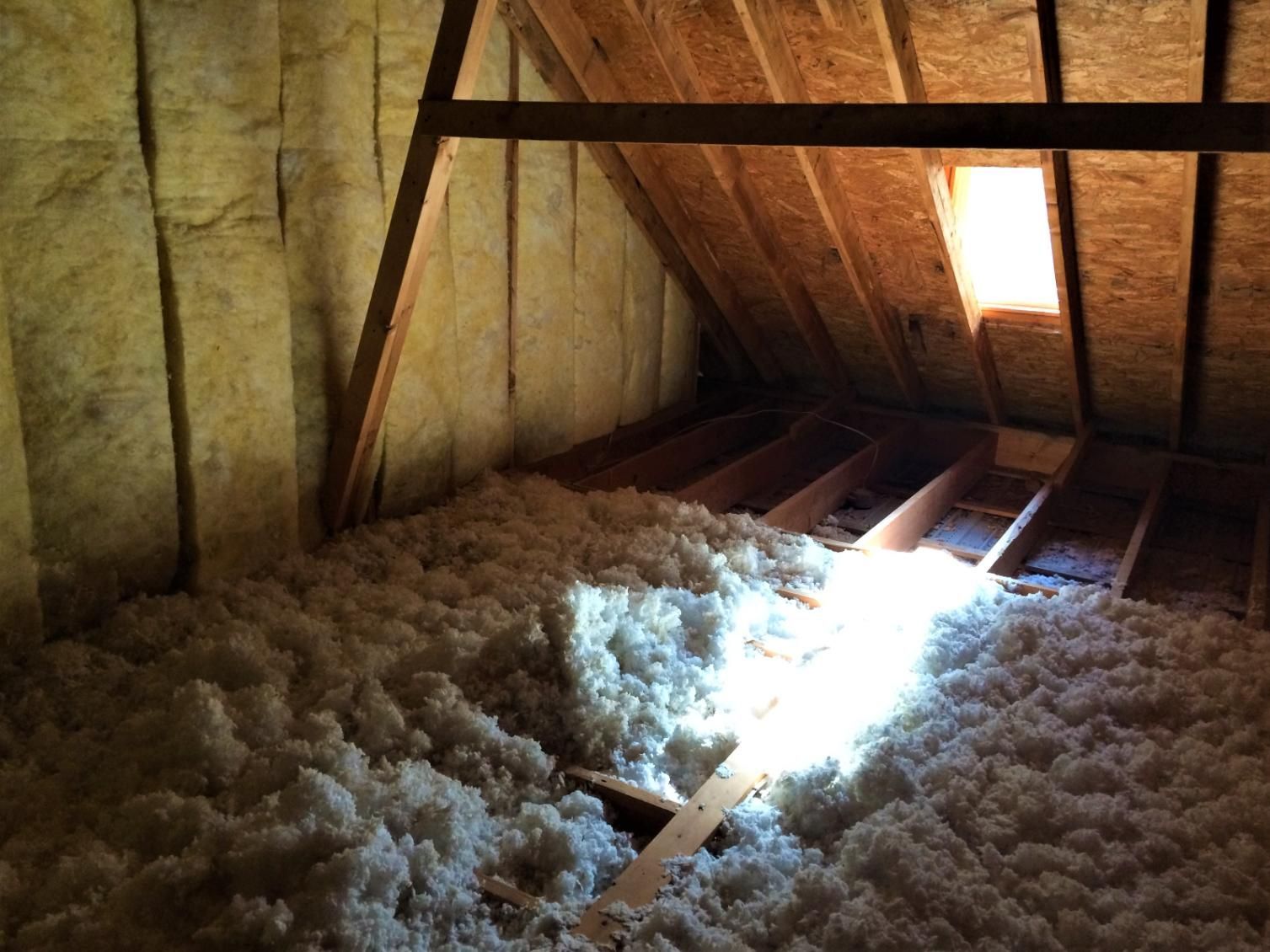 Attic interior filled with fluffy, white insulation. Wooden beams and unfinished walls.