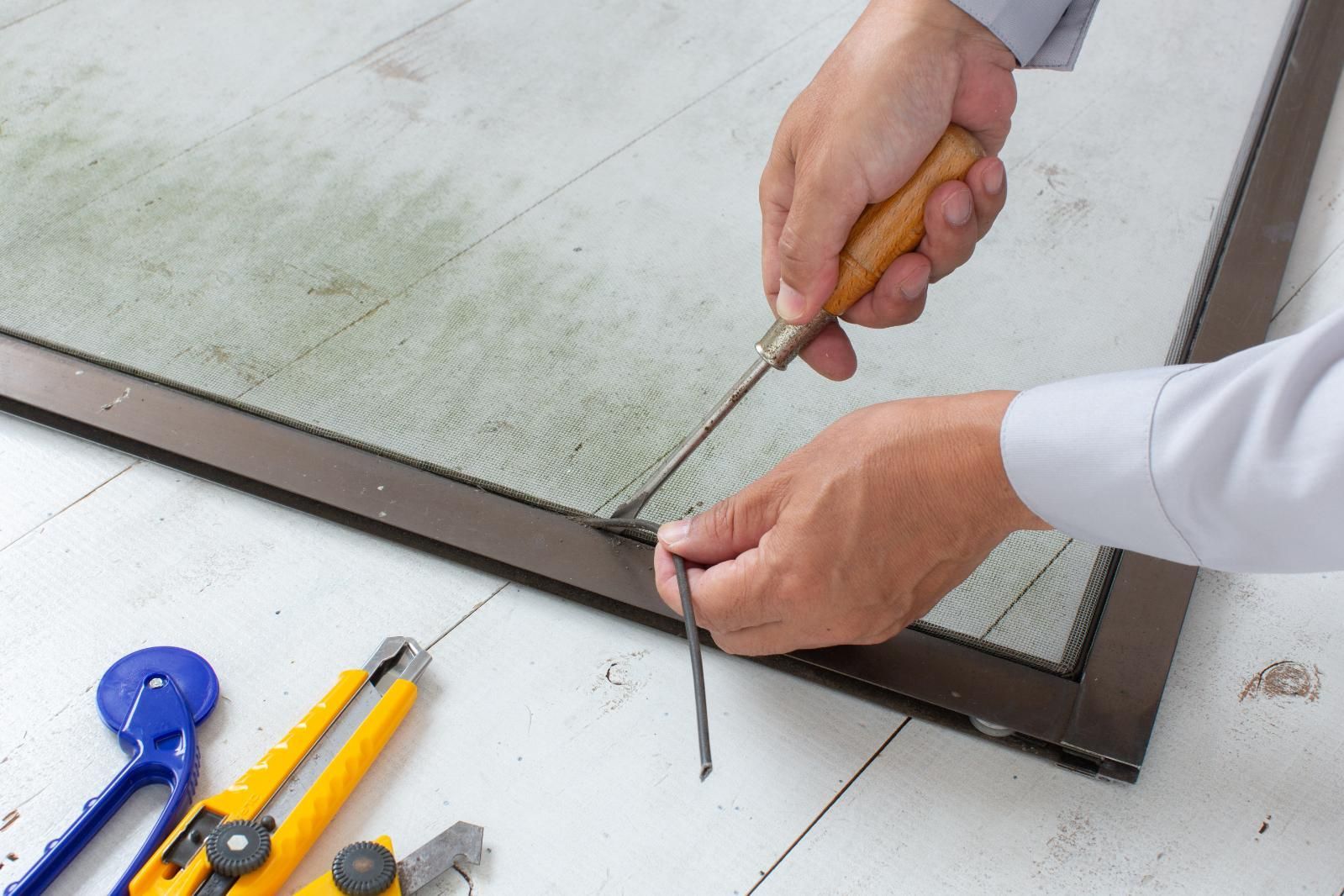 Person repairs a screen using a screwdriver and spline. Tools and a screen frame are in view.