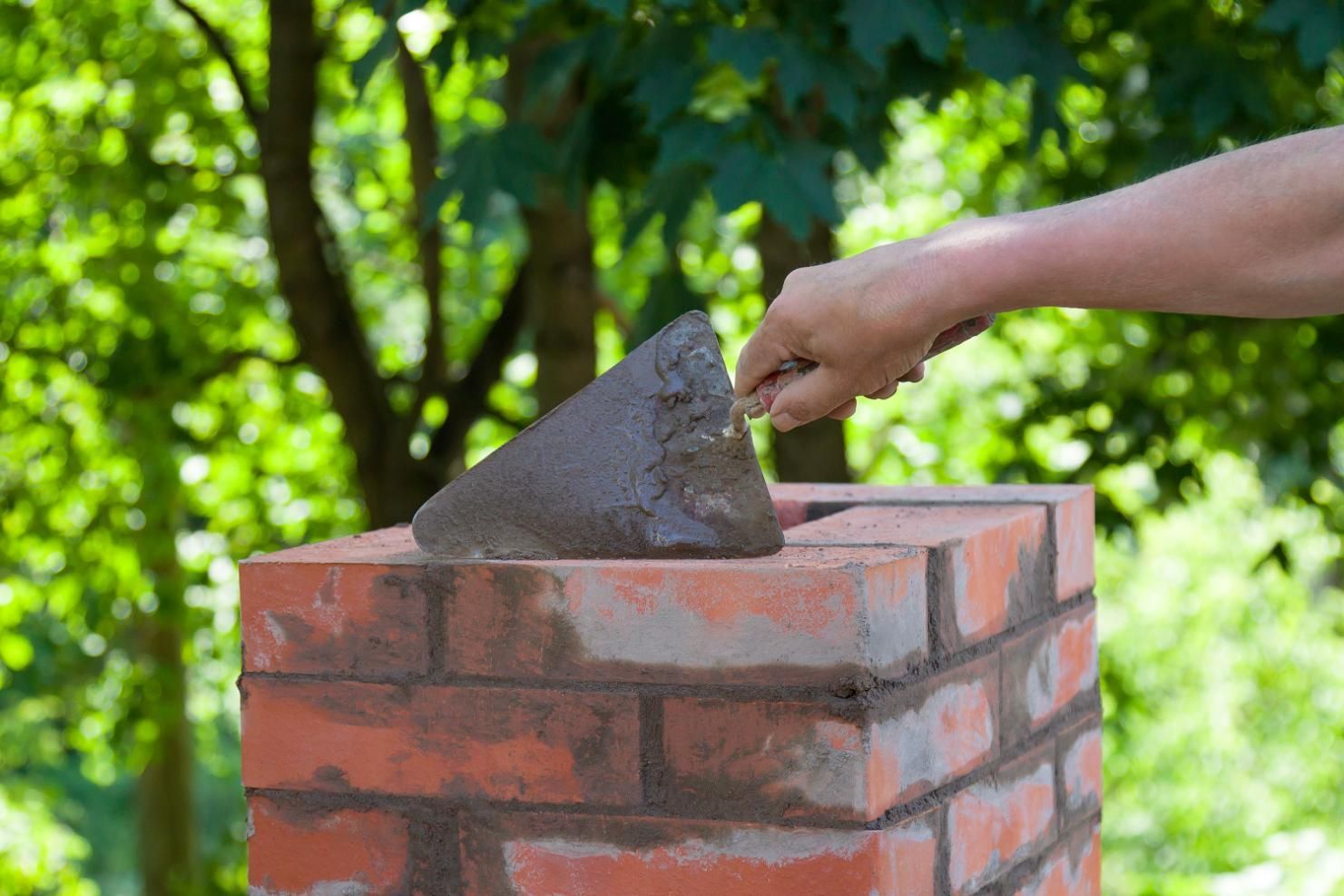 Hand using a trowel to apply mortar on brick chimney in outdoor setting.