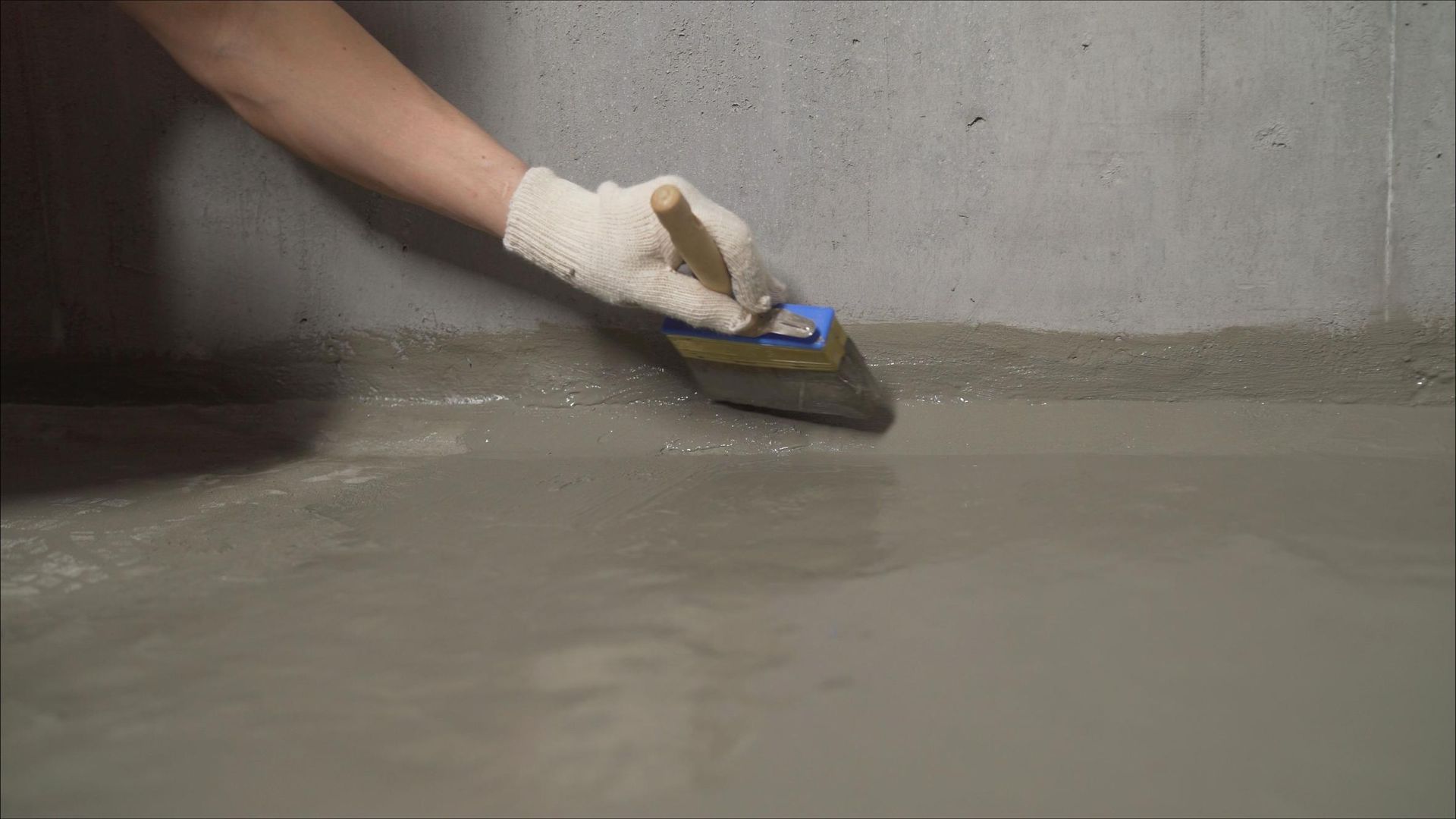 Person wearing a glove spreading concrete along a wall's edge with a trowel.