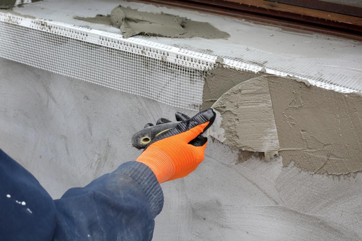 Person in orange gloves applying gray stucco with a trowel to a wall with metal mesh.