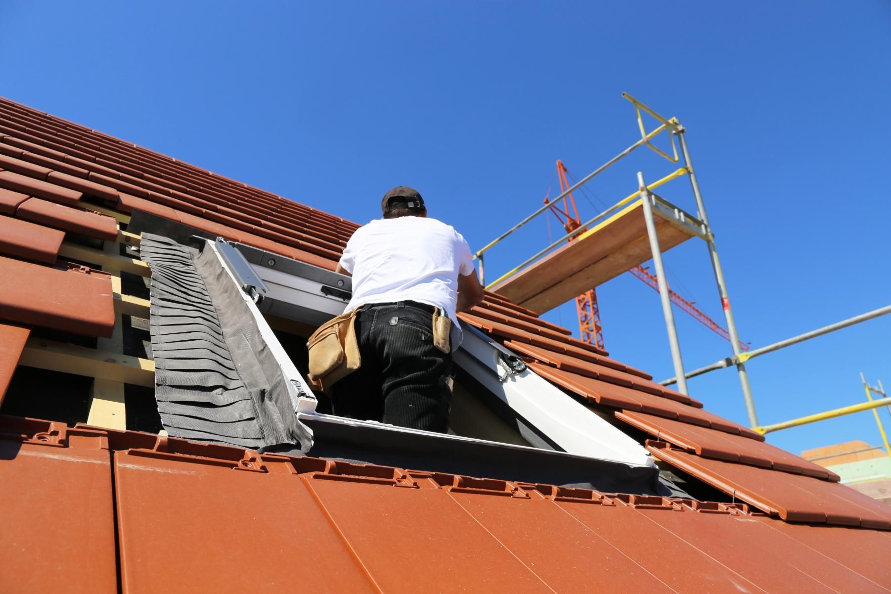 Roofer installing a skylight on a terracotta tile roof, with scaffolding in background.