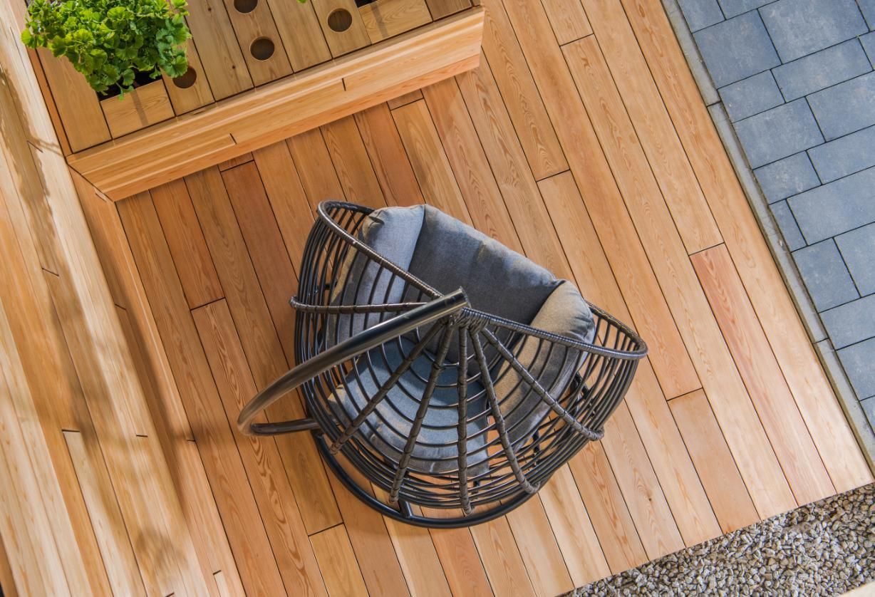 Wicker chair with cushions on a wooden deck, next to a planter and a paved area.