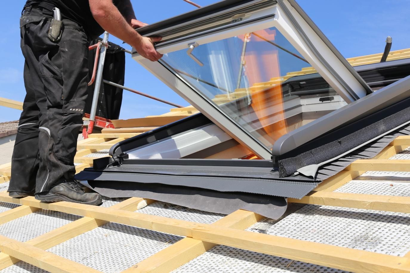 Person installs a skylight on a roof. Blue sky in the background.