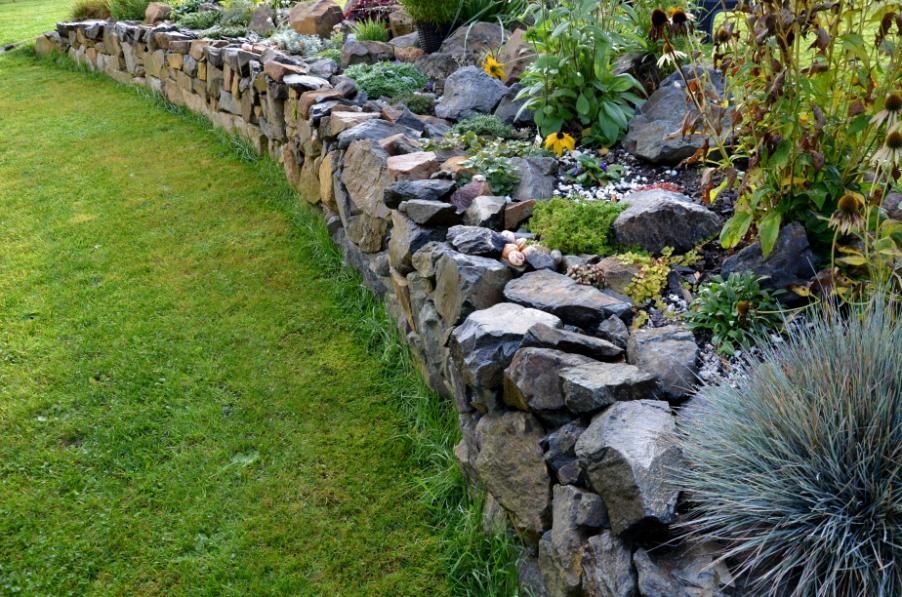 Stone retaining wall borders a garden bed filled with plants, beside a green lawn.