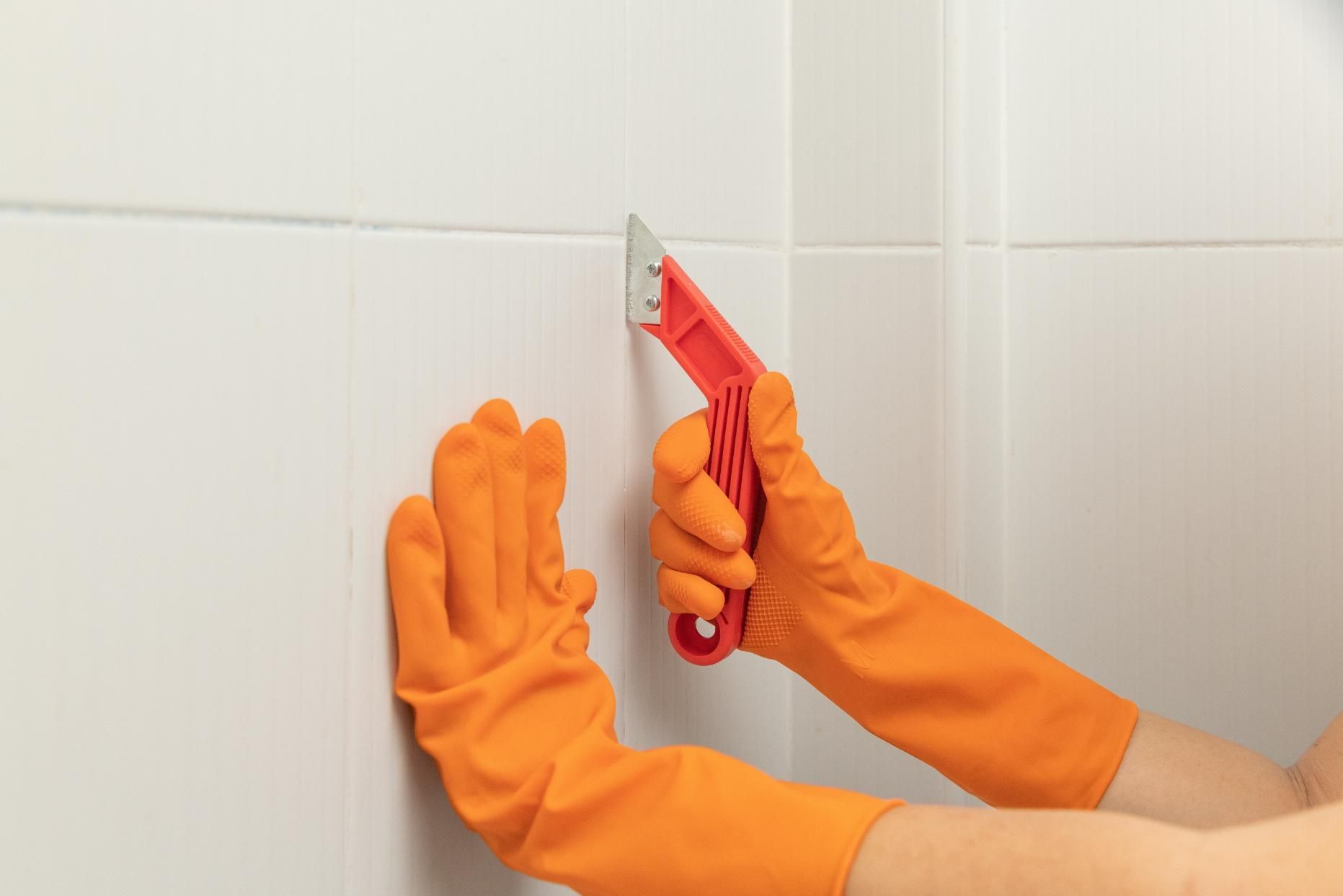 Hands in orange gloves using a red tool to remove caulk from white bathroom tile.