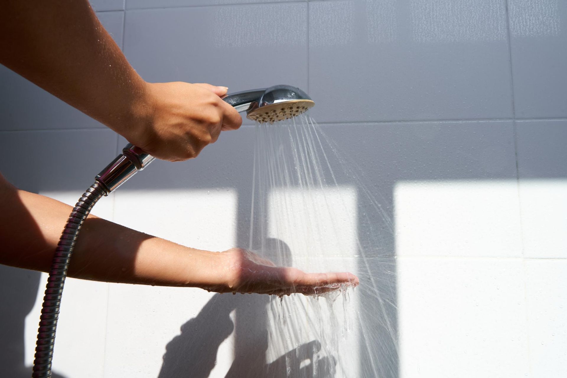 Person testing the water temperature from a shower head with their hand. White tiled wall background.