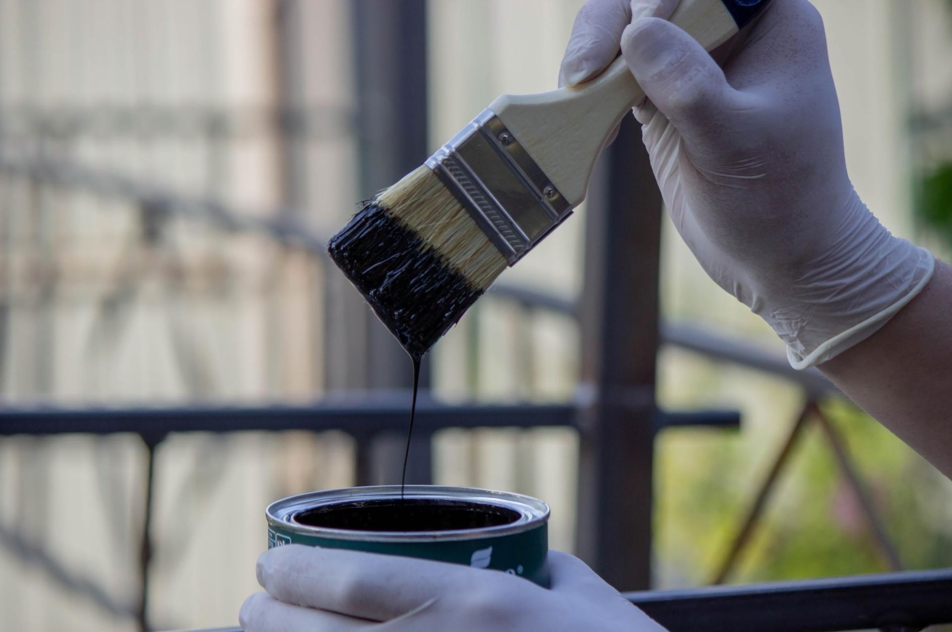 Person in gloves dipping a paintbrush into a can of black paint outside.
