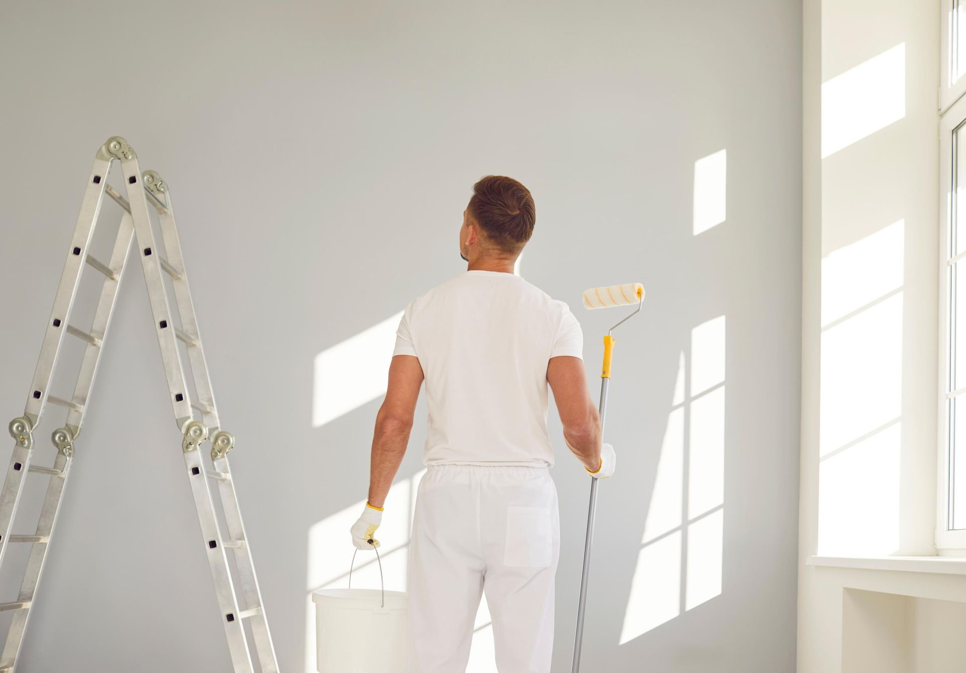 Person painting a wall, holding a paint roller, with a ladder and window present.