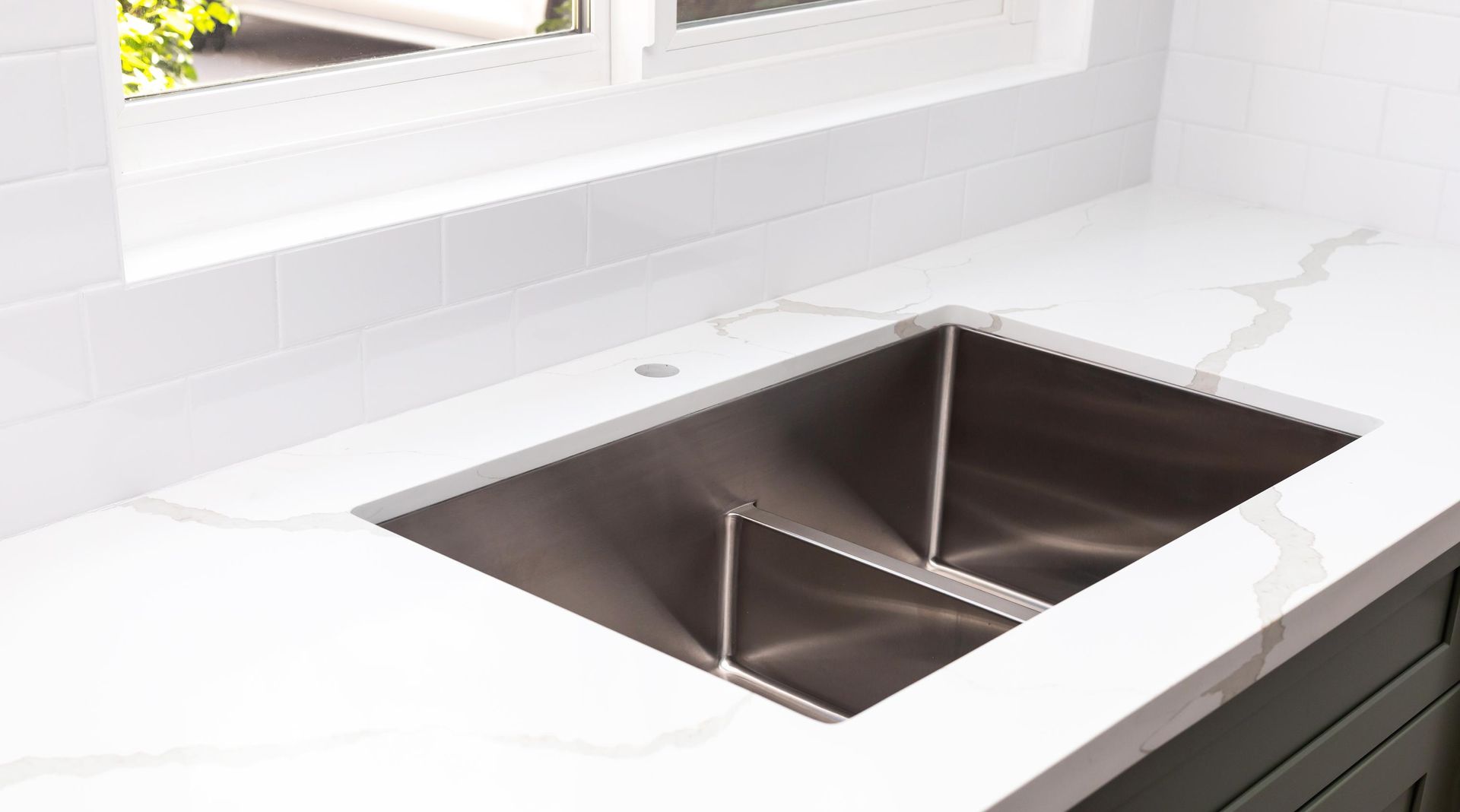 Stainless steel kitchen sink in a white countertop with a window in the background.