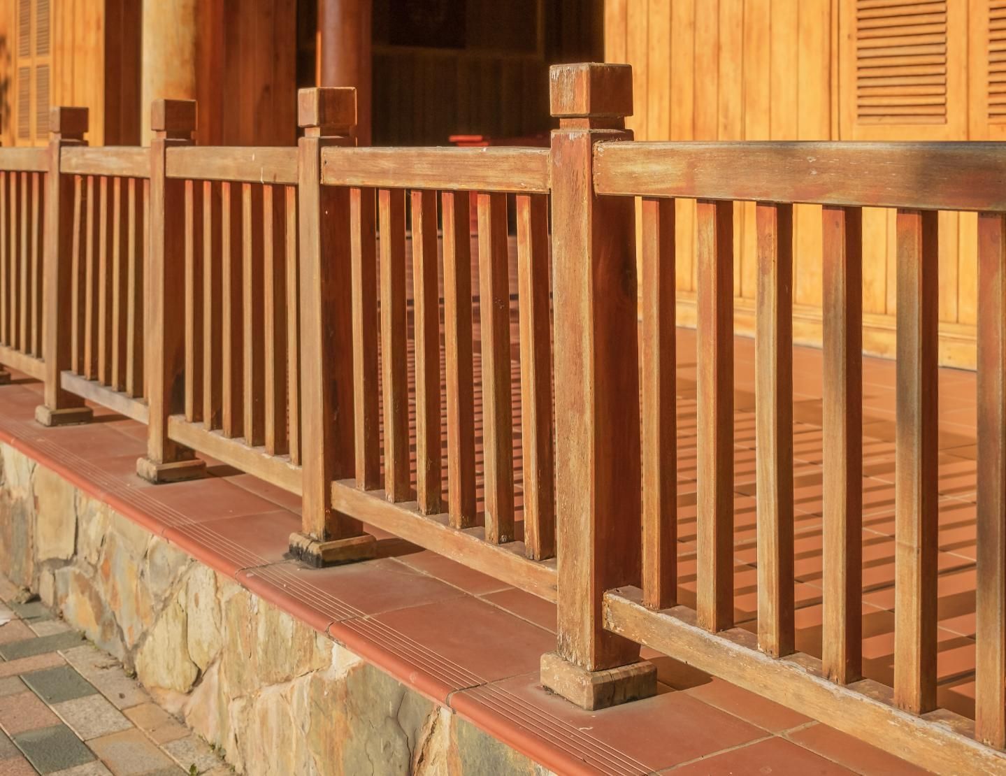Wooden railing along a building's porch, with vertical balusters. Light brown, with a stone and brick base.