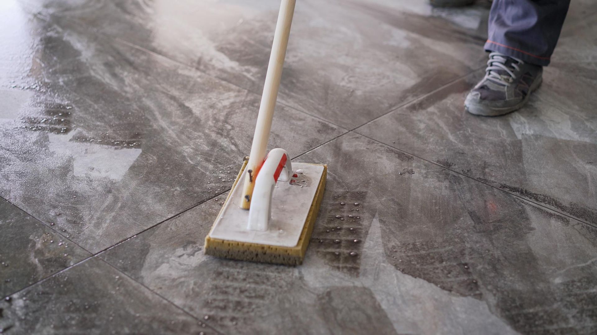 Person mopping a wet, patterned tile floor with a long-handled sponge mop.