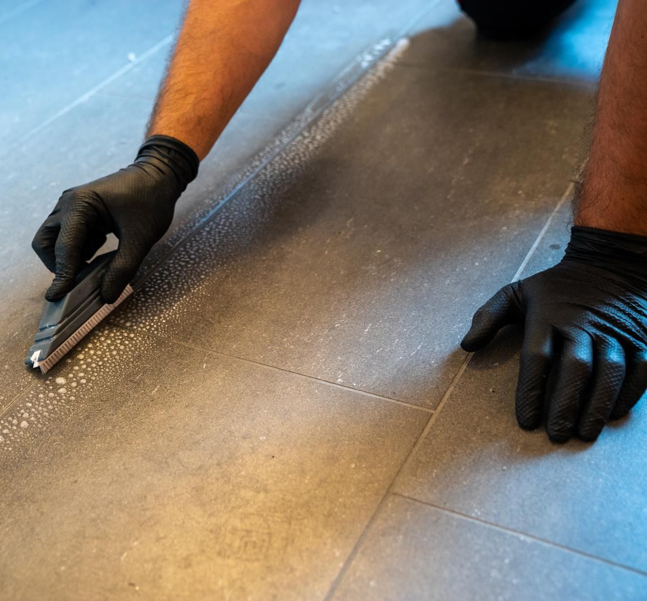 Person in black gloves, using a tool to apply something to gray tiled floor.