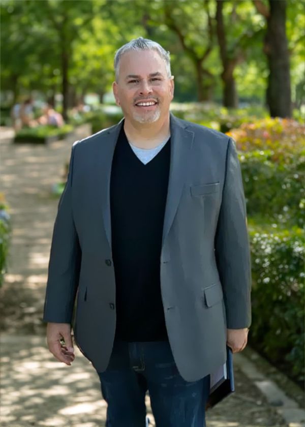 A man in a suit and black sweater is standing in a park.