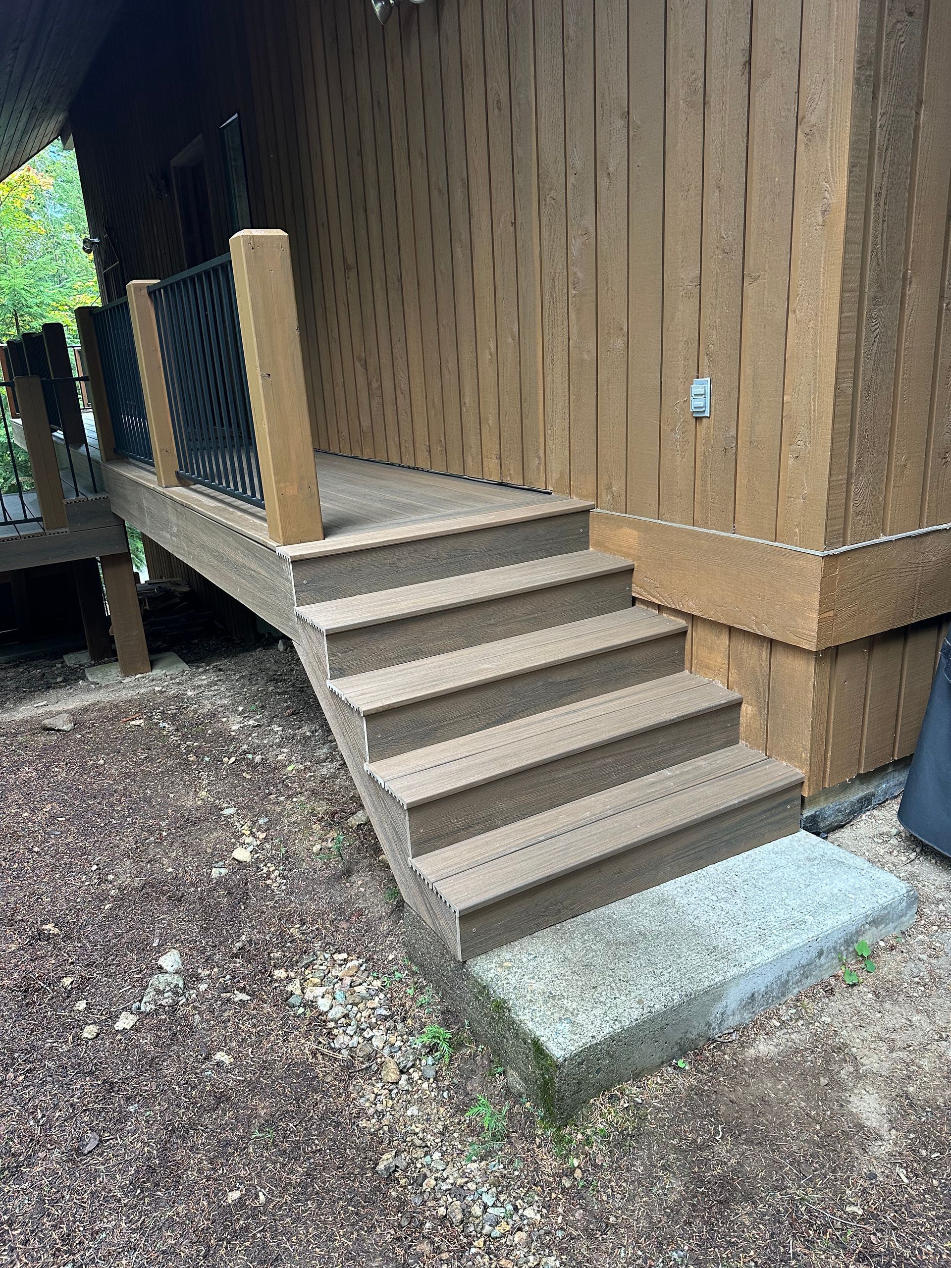 Brown composite deck stairs leading up to a wooden building. A small, light gray concrete pad is at the base.