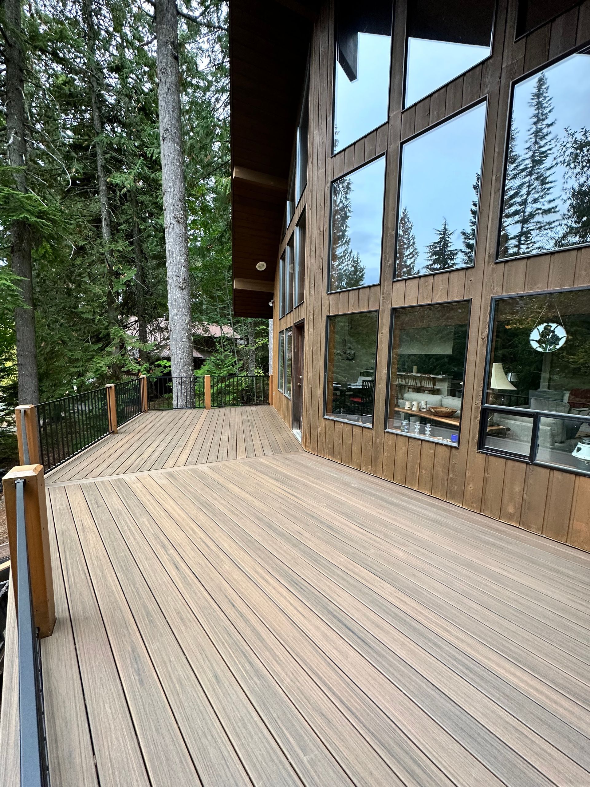 A wooden deck extends alongside a building with large windows reflecting the sky and surrounding trees.