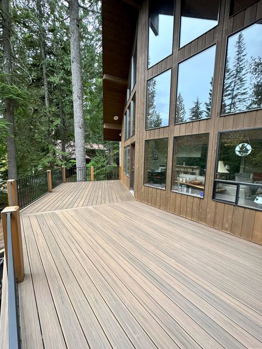 Wooden deck with handrails next to a building with large windows. Trees are visible behind the deck and windows.