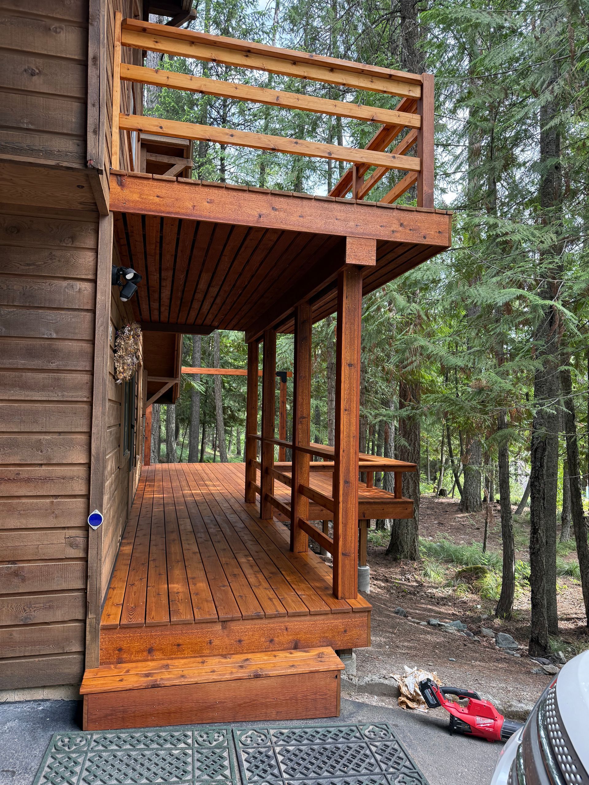 Wooden deck and railing attached to a cabin in a forest setting. Brown wood with a red lawnmower at the base of the deck.