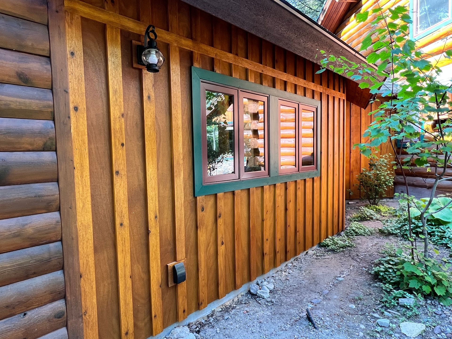 Exterior of a log cabin with vertical wood siding and a window framed in green. A small garden with plants is next to the building.