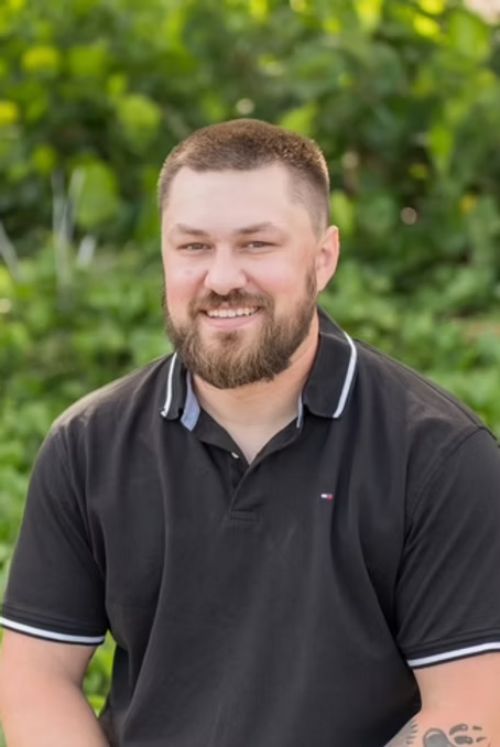 Man with a beard and dark hair, wearing a black polo shirt, smiles outside against a blurred green background.