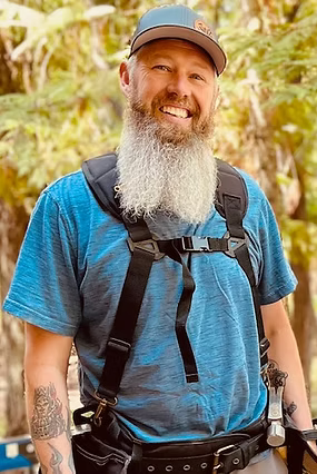 Man with a long gray beard and a blue cap smiling, wearing work gear outdoors with a wooden background.
