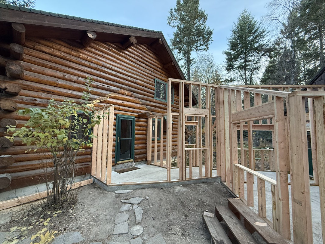 Framed walls being built onto the side of a wooden log cabin. The construction is outdoors, with trees and sky in the background.
