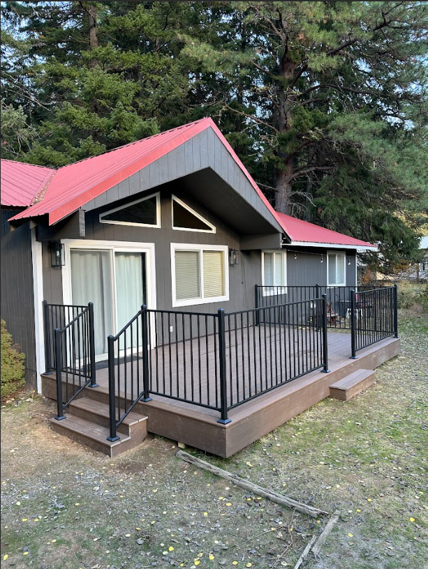 A gray cabin with a red roof and a black railed deck sits in a grassy area surrounded by trees.