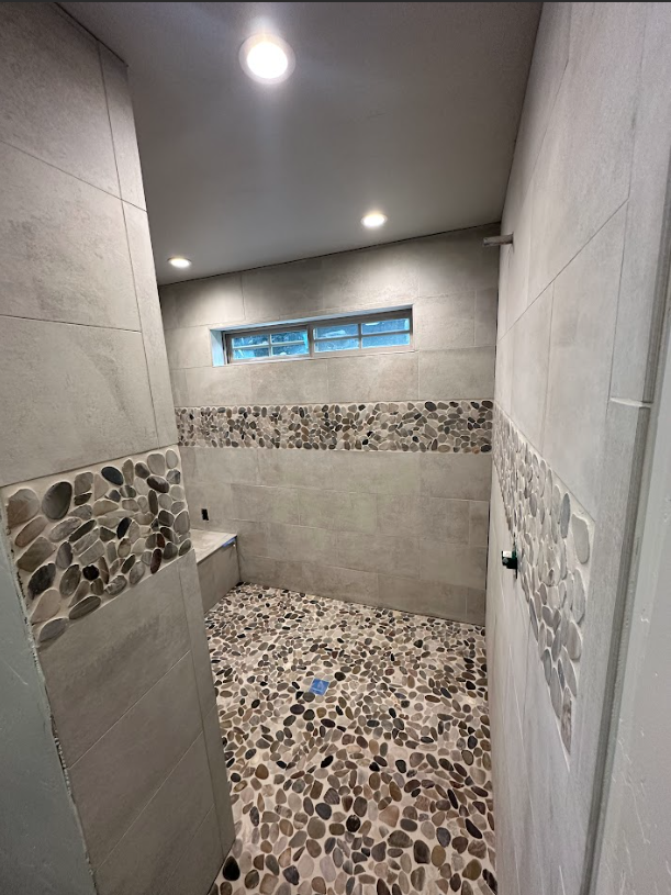 A modern shower with gray tile walls, pebble floor, a small window, and decorative stone accents.