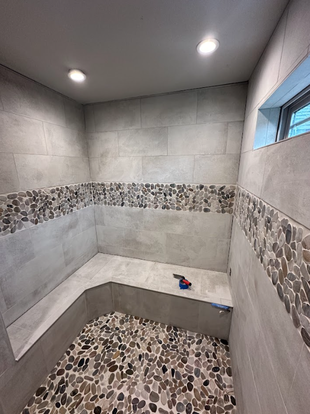 A newly tiled shower with pebble flooring, gray walls, a built-in bench, and a strip of stone tile.