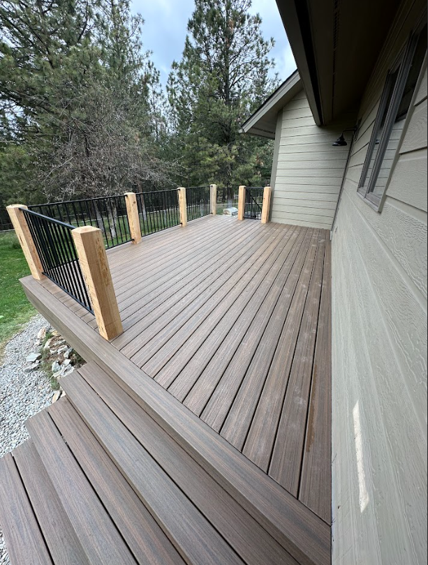 A deck with gray composite decking boards, stairs, and black metal railings. The deck has wooden posts and is next to a house with green siding.