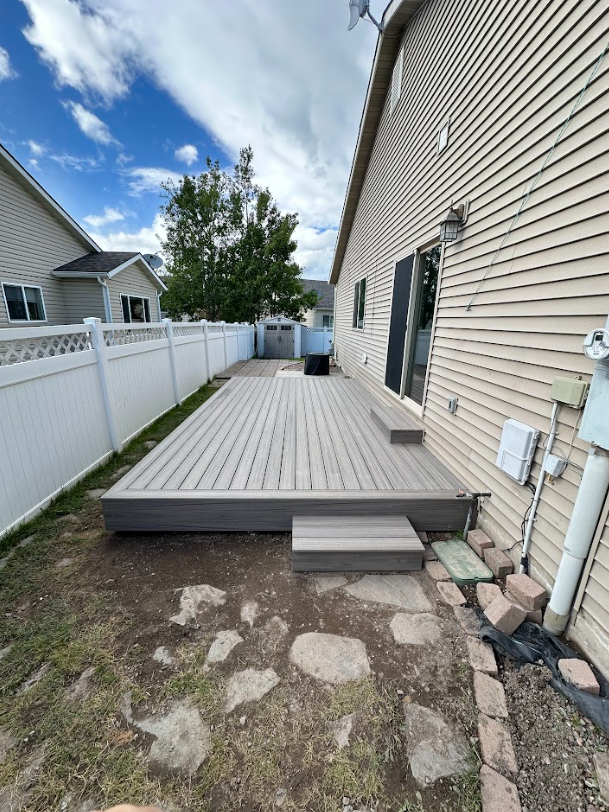 A composite deck with steps, next to a light-colored house. A white fence and backyard complete the setting on a partly cloudy day.