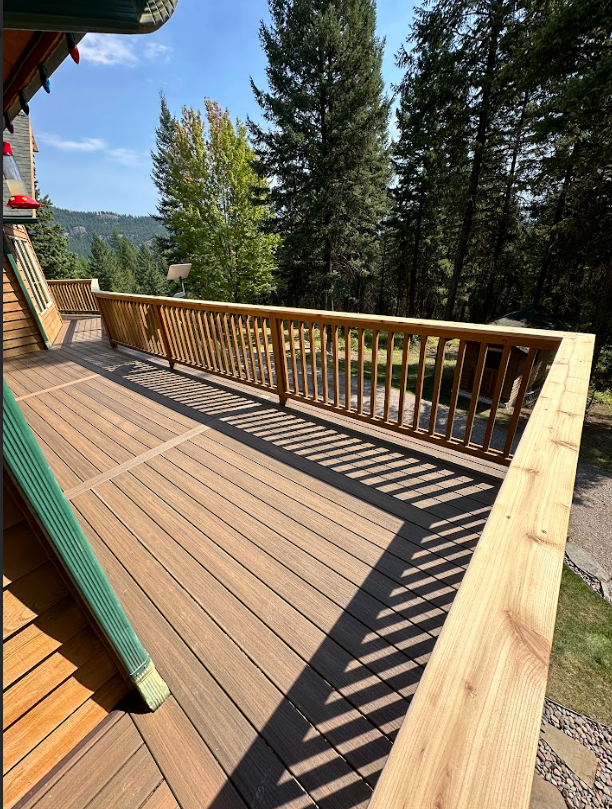 Wooden deck with brown composite decking, wooden railing, and a green-trimmed facade, overlooking a sunny forest.
