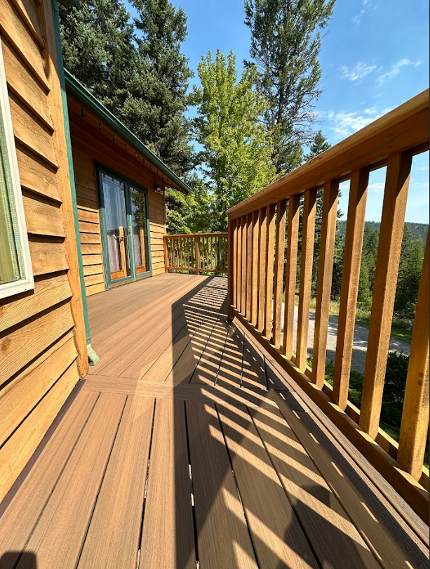 Wooden deck with railing and view of trees, adjacent to a wood-sided house under a sunny sky.