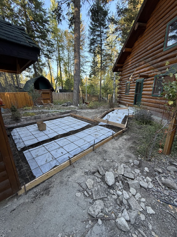 Construction site with wooden forms and wire mesh laid out for concrete, next to a log cabin. The setting is outdoors, in a backyard.