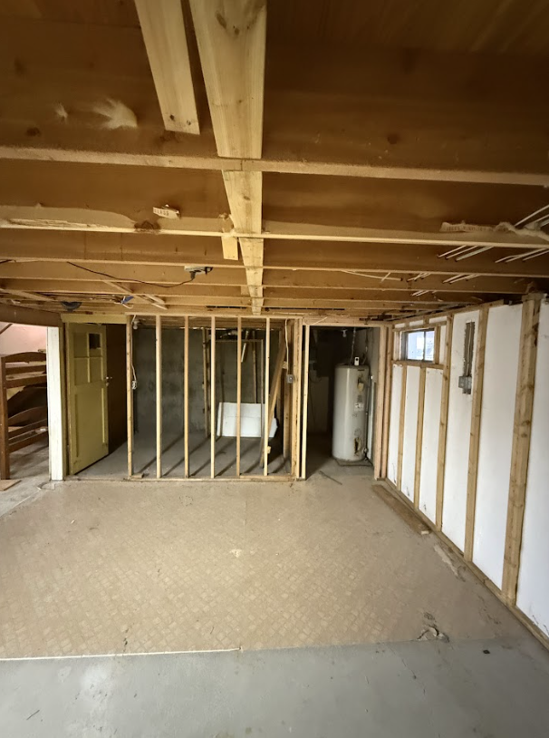 Unfinished basement with exposed wooden beams, partially framed walls, a water heater, and a concrete floor.