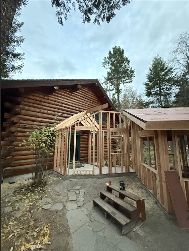 Construction of a wooden framed extension next to a log cabin. The extension is in progress, with walls and roof supports visible. Overcast day.