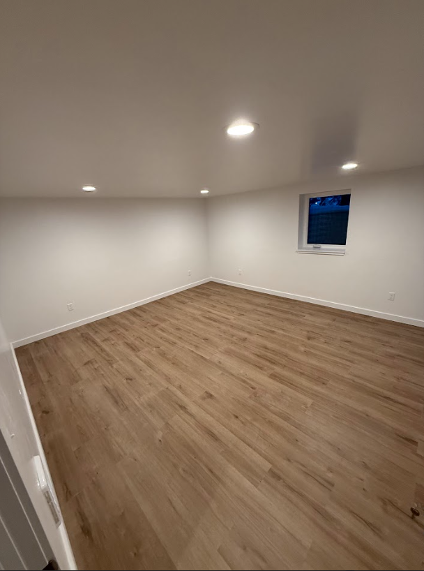 Empty room with wood-look flooring and white walls and ceiling. Recessed lighting and a small window are visible.