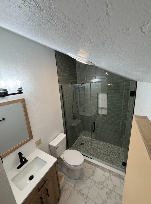Small bathroom with a shower, toilet, and vanity. Shower has glass doors and stone floor, with green tiled walls.