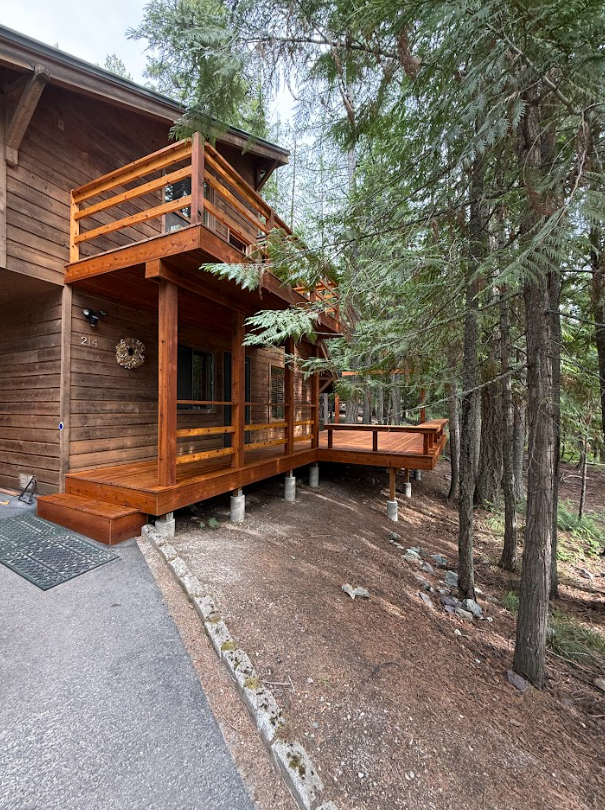 Wooden cabin with two-story deck in a forest setting. The deck is stained brown and supported by posts on a slope.